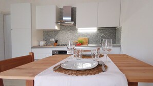 Une table à manger dressée avec des assiettes, des verres et des couverts, une chaise en bois, et en arrière-plan, une cuisine avec un plan de travail décoré et des armoires blanches.