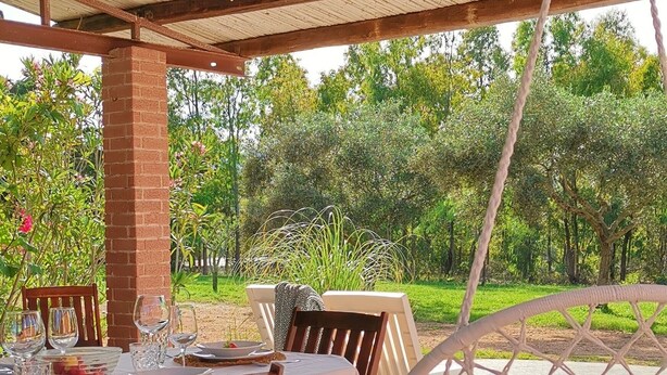 Un espace de restauration en plein air doté d’une terrasse couverte, de chaises en bois, d’un luminaire suspendu et offrant une vue sur un jardin.