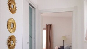 Un couloir avec une porte donnant sur une pièce où se trouvent un lit et une lampe. Trois éléments décoratifs ornent le mur.
