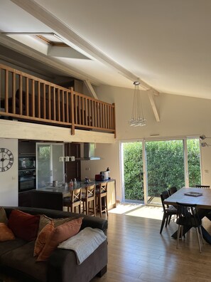 Un vaste espace de vie doté d’un haut plafond, un balcon avec des balustrades en bois, une cuisine équipée d’appareils modernes et un coin repas avec une grande table et des chaises.