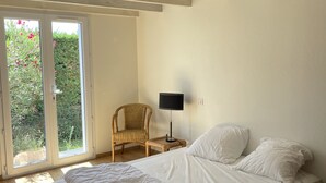 A bedroom with a large bed, a chair, a nightstand with a lamp, and a view through a sliding door to a garden.