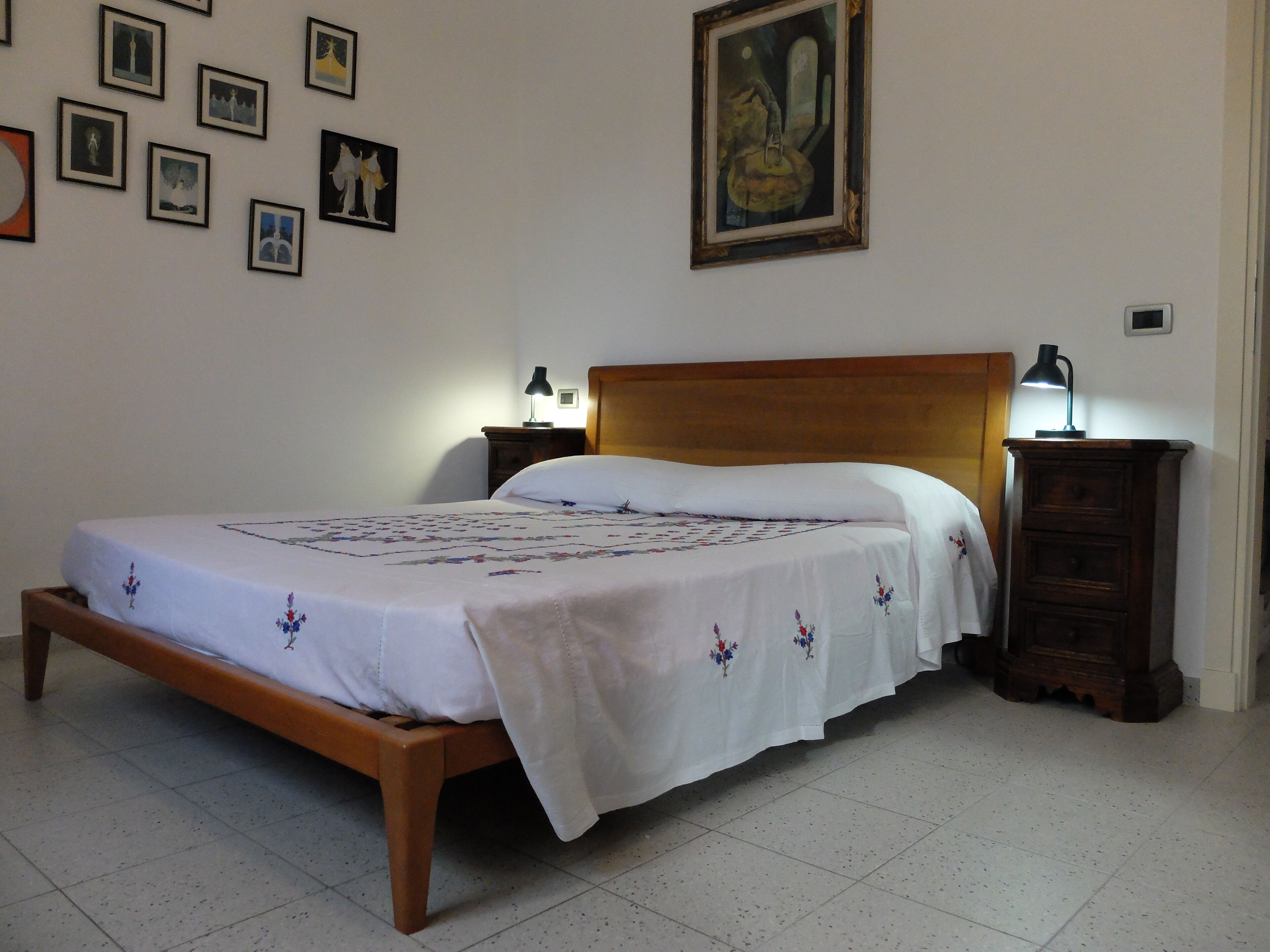 A bedroom with a wooden bed, white bedding, a nightstand with a lamp, and a wall adorned with framed pictures.
