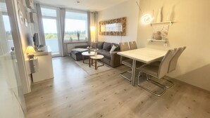 Un salon moderne comprenant un canapé, deux fauteuils, une petite table et une table à manger avec des chaises. On y trouve une fenêtre avec des rideaux, un radiateur et un luminaire fixé au mur.