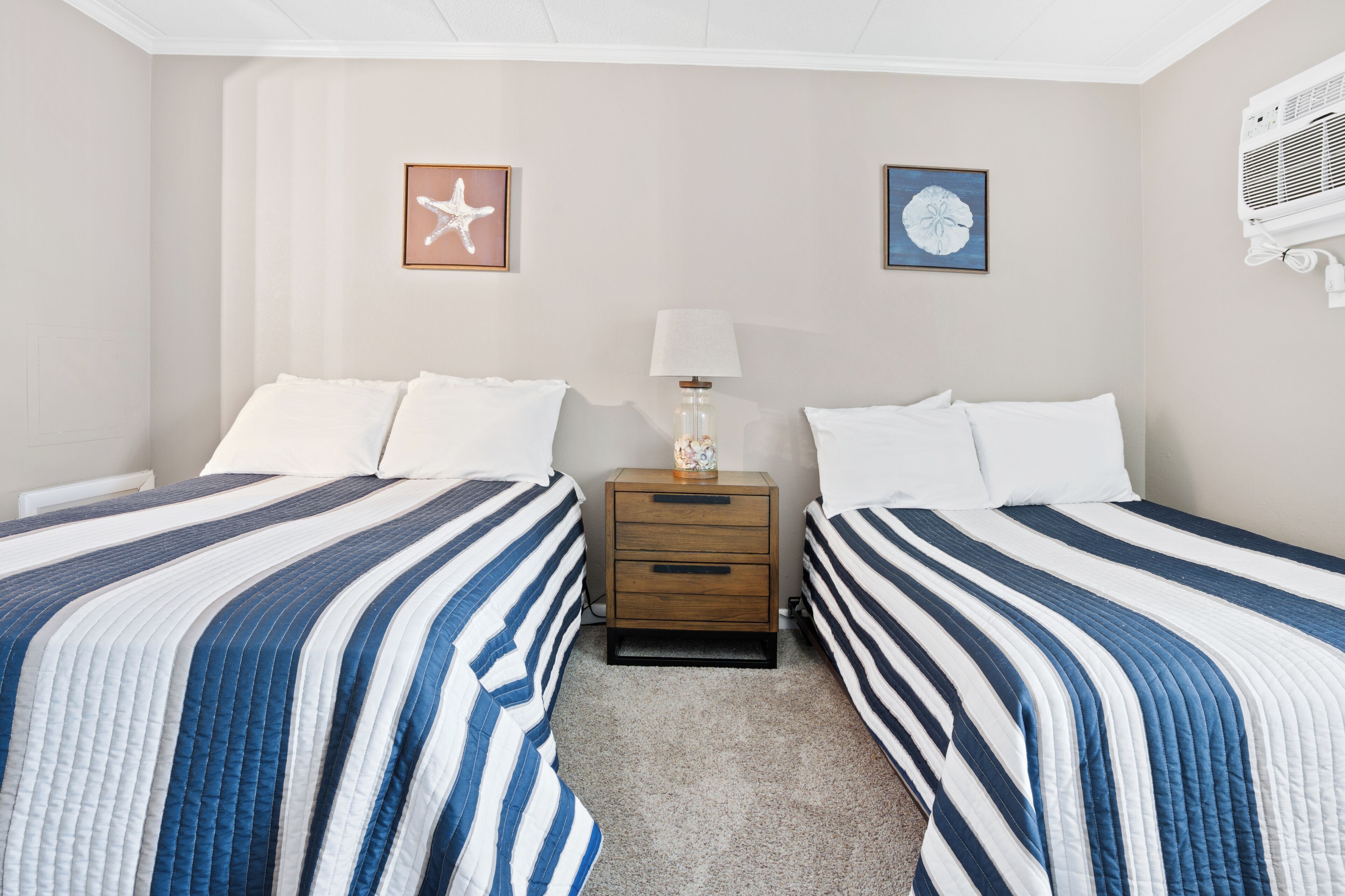 A hotel room with two beds, a nightstand, a lamp, and two wall art pieces.