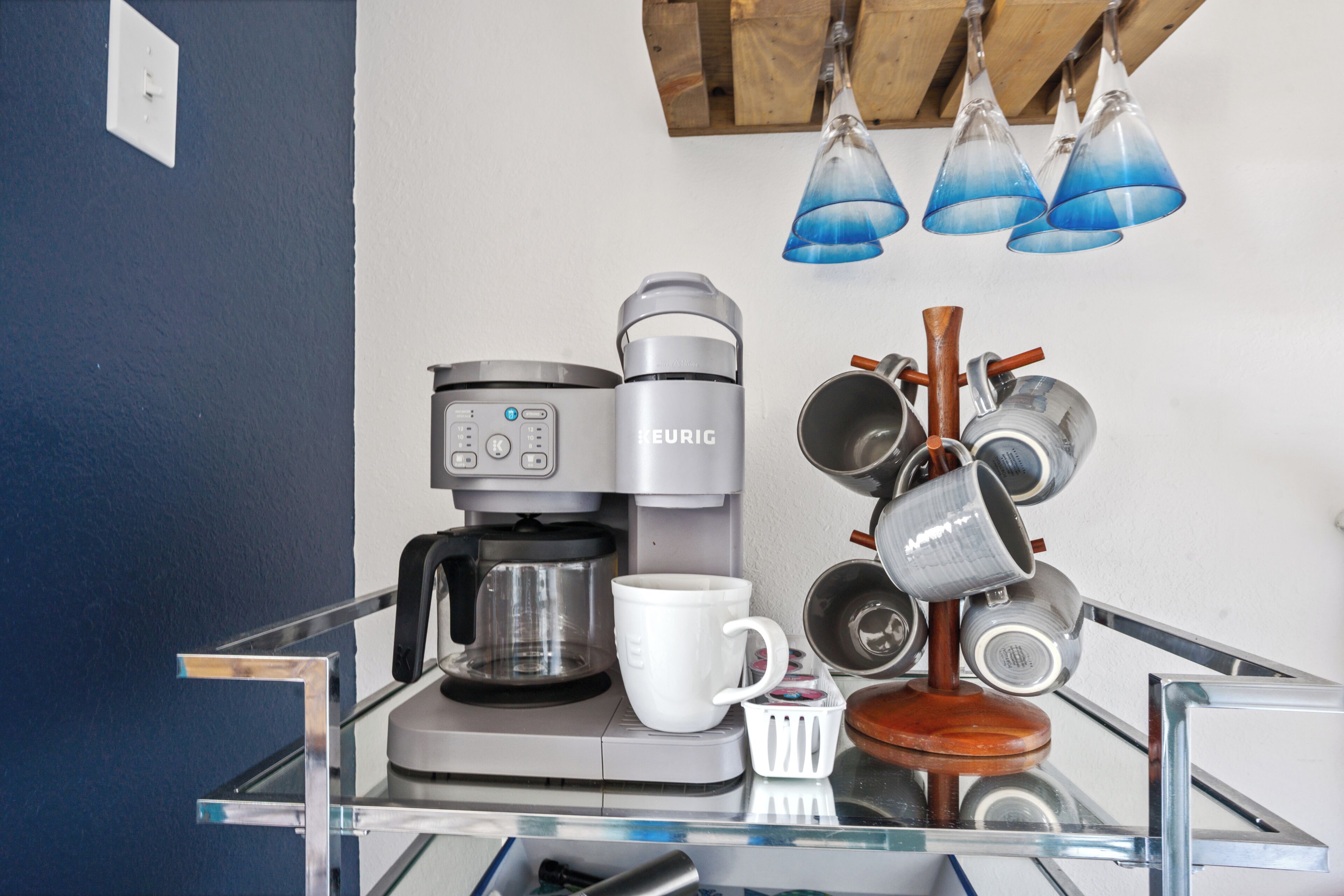 Een keuken met een koffiezetapparaat, een witte mok en een kruidenrek met meerdere pannen.