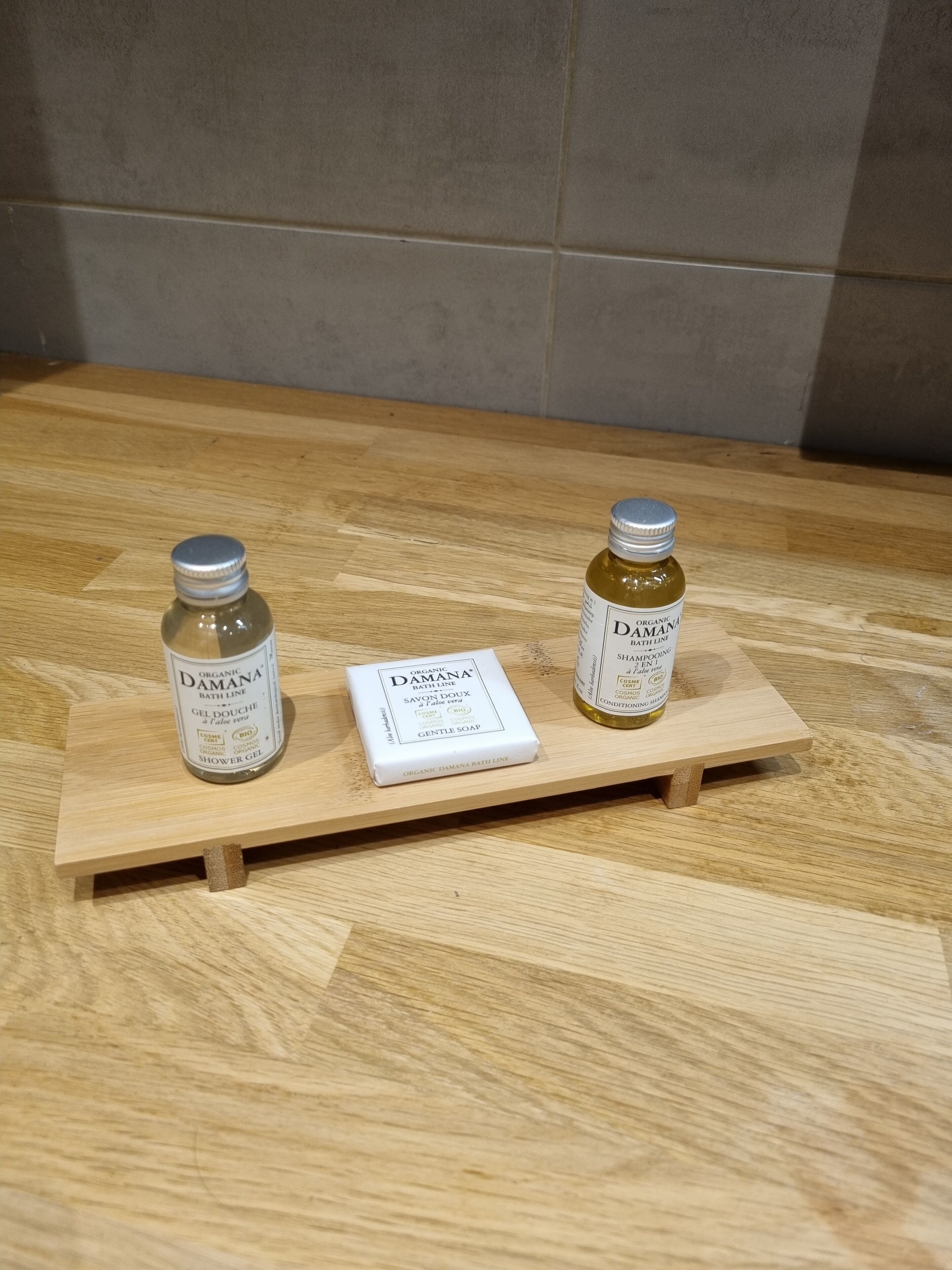 A wooden tray with two bottles of Organic Damana Bath Line and a box of Organic Damana Gentle Soap on a wooden floor.