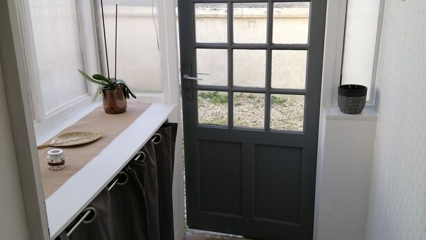 A room with a black door, a window with blinds, a potted plant, and a small shelf with a container. - Charming 70m2 town house 15kms from the sea and 4 kms from Caen. Parking (Giberville)