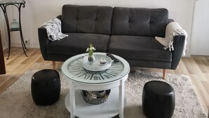 A living room with a dark grey sofa, a glass coffee table with Roman numerals, a white rug, and three framed artworks on the wall. - Charming 70m2 town house 15kms from the sea and 4 kms from Caen. Parking (Giberville)