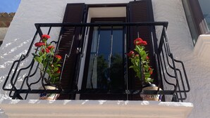 Un balcon avec un mur blanc et un toit recouvert de tuiles. On y trouve deux plantes en pot, ornées de fleurs rouges.