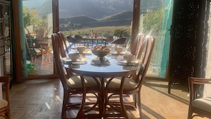 Un coin repas avec une table dressée pour quatre personnes, entourée de chaises, et offrant une vue sur les montagnes à travers de grandes portes vitrées.