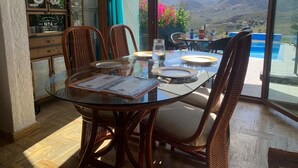 Un coin repas avec une table en verre, des chaises en osier et une vue sur les montagnes, visible à travers de grandes fenêtres.