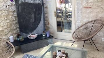 Een kamer met een stenen muur, een groot zwart kunstwerk, rieten stoelen, een glazen tafel met decoratieve objecten en een deur met uitzicht op buiten.