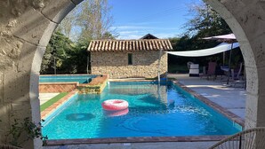 A swimming pool with a stone house in the background, surrounded by outdoor furniture and greenery.