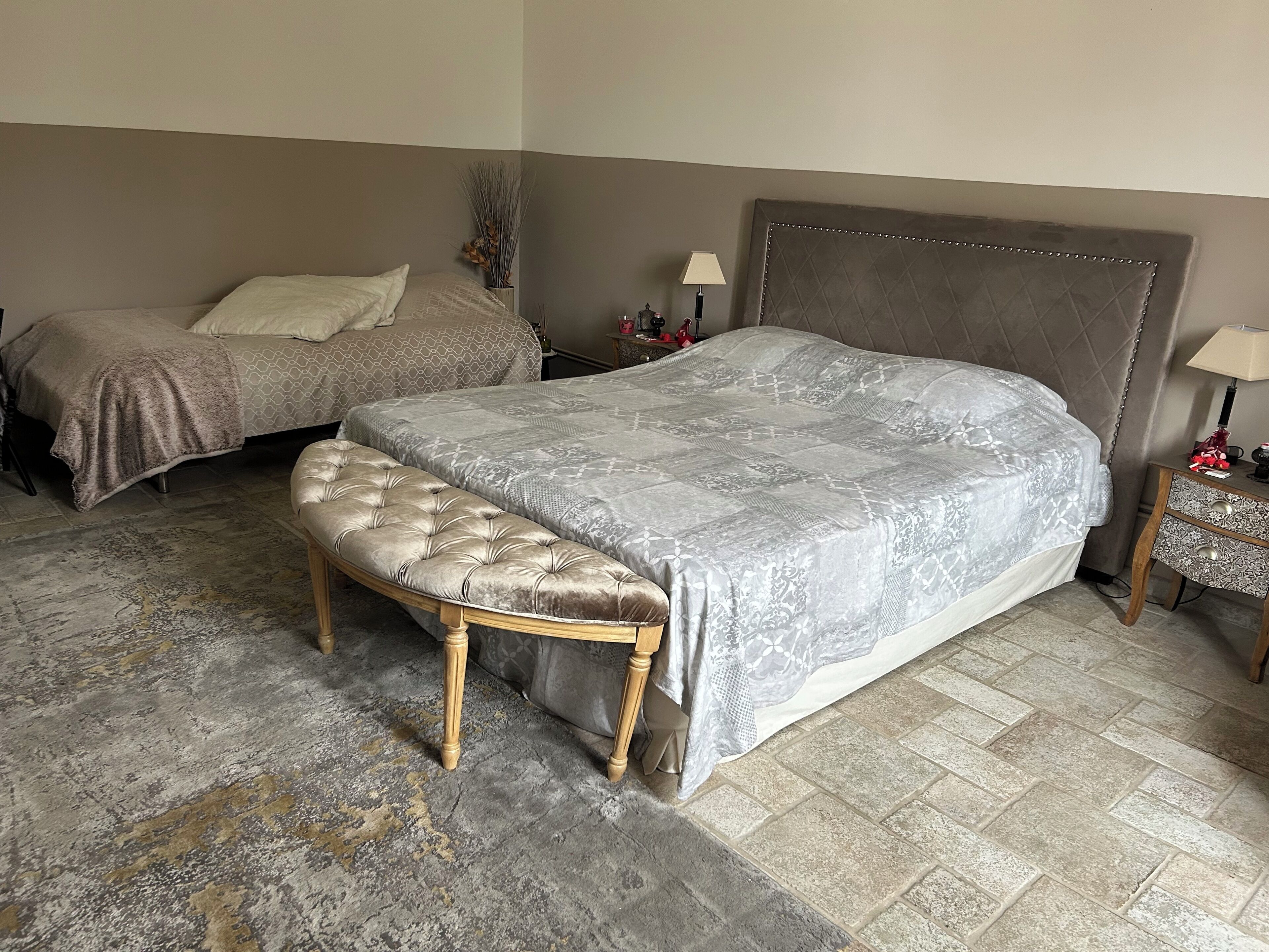 Un dormitorio con una cama grande, una cama más pequeña, un banco, dos mesitas de noche y una planta decorativa.