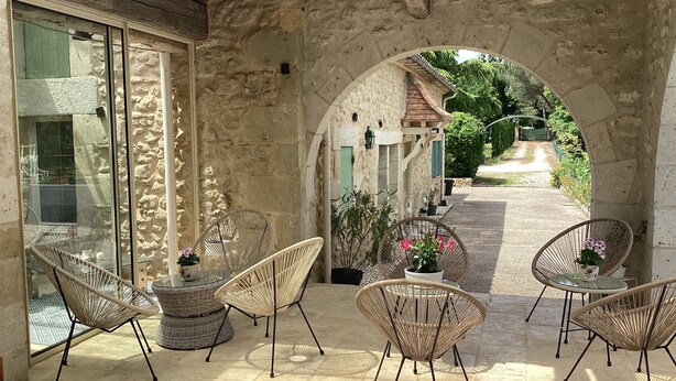 A covered patio with wicker chairs and potted plants, stone walls, and a view of a street.
