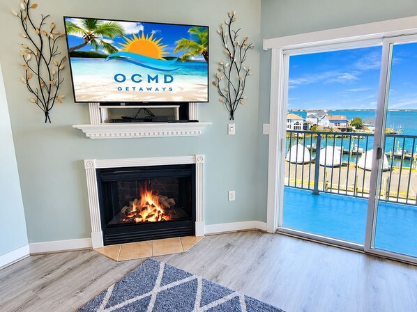 Una habitación con chimenea, un televisor mostrando 'OCMD GETAWAYS' y vista al mar desde un balcón.