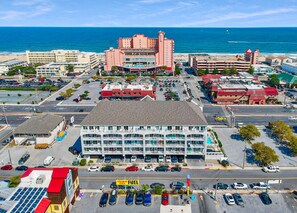 An aerial view of a coastal city with a prominent hotel featuring multiple floors and balconies, a parking lot, and a view of the ocean. - Luxury 3 Bedroom Bayview Condo with Pool - 22nd St! Overlooking Bahia Marina and Fish Tales! Mariner's Watch #304 (Ocean City)