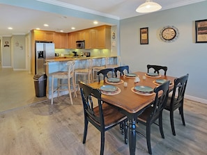 A dining area with a wooden table set for six, surrounded by chairs. A kitchen with wooden cabinets and a refrigerator is visible in the background. - Luxury 3 Bedroom Bayview Condo with Pool - 22nd St! Overlooking Bahia Marina and Fish Tales! Mariner's Watch #304 (Ocean City)