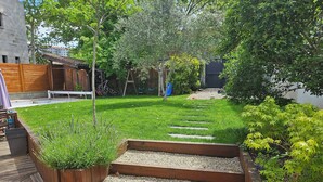 Un giardino con un patio in legno, un sentiero ghiaioso e diverse piante.