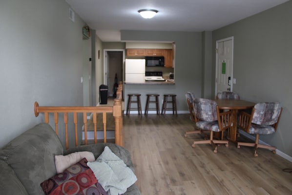 Una sala de estar con un sofá gris, una mesa de comedor de madera con cuatro sillas y una cocina con armarios de madera y un microondas.