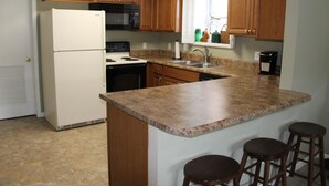 A kitchen with wooden cabinets, a white refrigerator, a stove, a sink, and three bar stools. - Seaworthy at Wheelhouse Lake Ozark MO (Lake Ozark)