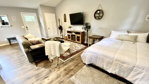 A spacious bedroom with a large bed, a sitting area with a sofa and chairs, a TV mounted on the wall, and a wooden floor. - TC TWO OAKS Studio / Downtown / Clean & Spacious (Traverse City)