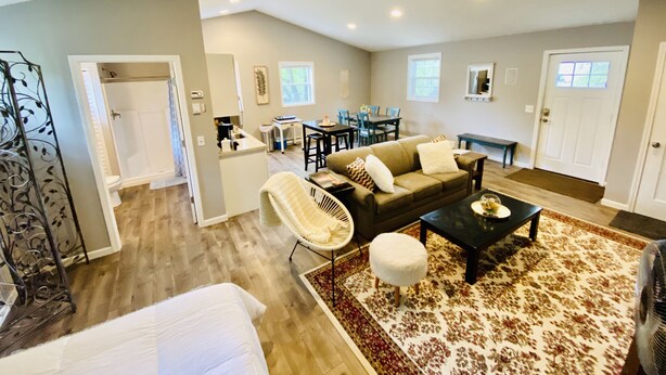 A spacious living area with a sofa, coffee table, and a dining table with chairs. There is a separate dining area with a window, and a bathroom with a shower and toilet visible. - TC TWO OAKS Studio / Downtown / Clean & Spacious (Traverse City)