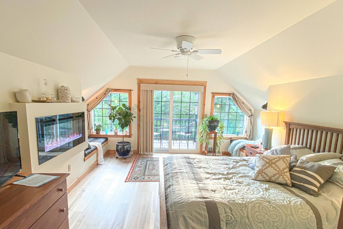 A bedroom with a bed, a desk, a TV, a ceiling fan, and large windows.