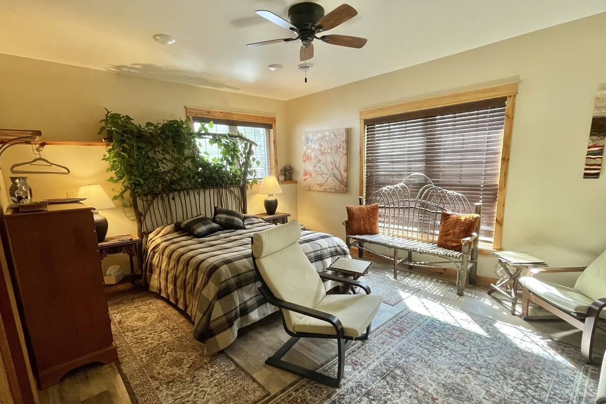 A bedroom with a bed, a chair, a desk, a plant, and a window with blinds.