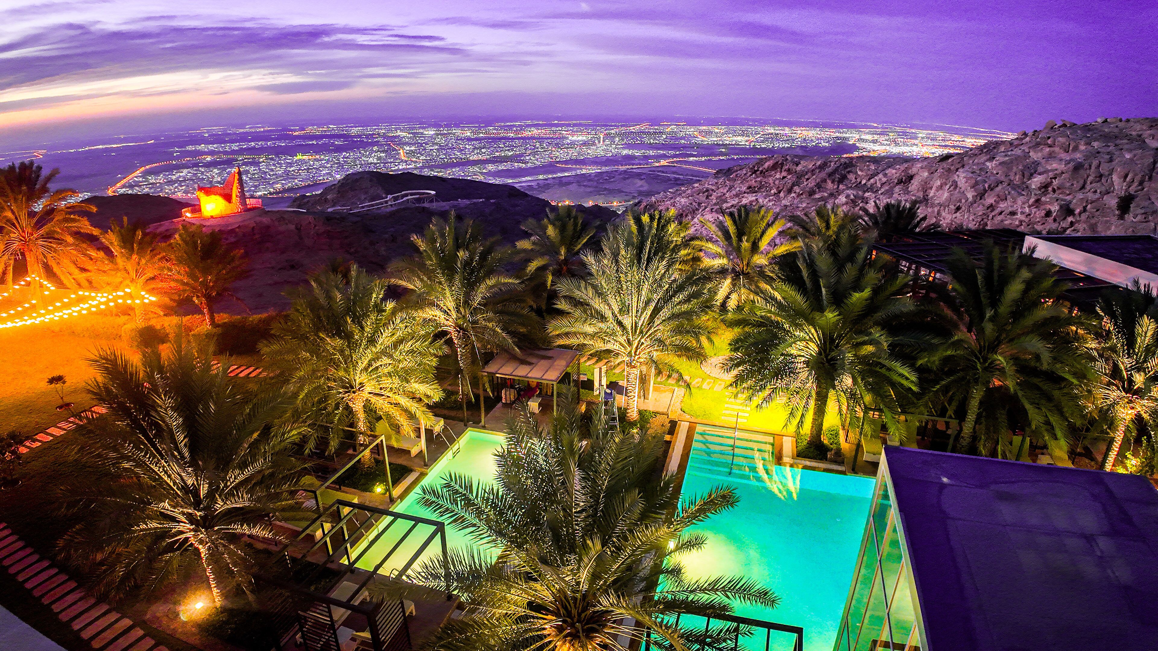 A resort with a swimming pool, palm trees, and a view of a cityscape at night.