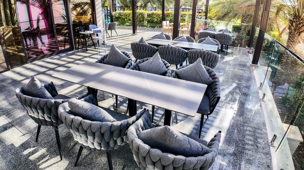 A modern outdoor seating area with a central table and cushioned chairs, surrounded by glass railings and greenery. - Alberni Jabal Hafeet Hotel (Al Ain)