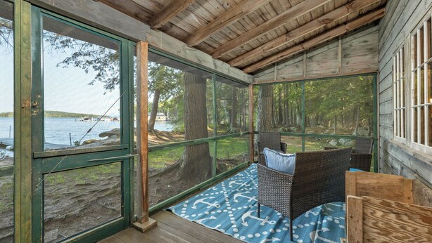 A wooden cabin with a large glass door and windows overlooking a lake. Inside, there is a wicker chair with cushions and a blue patterned rug on the floor.