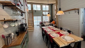 Et spiseområde med et langt bord dækket til fire personer, træbjælker i loftet og en trappe, der fører op til et højere niveau.