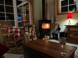 Un salon chaleureux avec une cheminée, un fauteuil à bascule en bois, une table avec une bouteille de vin et une fenêtre donnant sur la ville illuminée la nuit.