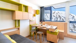 A modern hotel room with a bed, a desk with two stools, a small kitchen area, and a large window with a mountain view. - Home Club (Lavachet) by Interhome (Tignes)