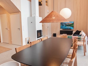 A modern living room with a large dark-colored dining table, wooden chairs, a television mounted on the wall, and a woven light fixture.