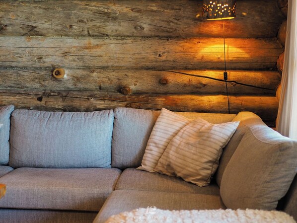 A log cabin interior with a sofa against a wooden wall, a hanging light fixture, and a window with curtains.