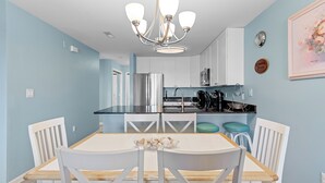 A kitchen with white cabinets, a stainless steel refrigerator, a microwave, and a sink. There is a dining table with white chairs and a chandelier above. - Baywatch II 102A at Heron Harbour by Seaside Vacations & Sales (Ocean City)