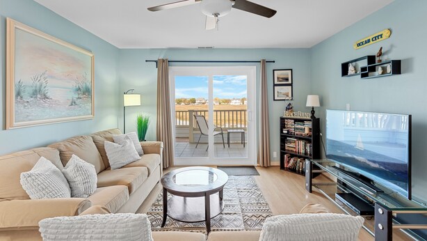 A living room with a beige sofa, a glass coffee table, a ceiling fan, a TV, and a bookshelf. - Baywatch II 102A at Heron Harbour by Seaside Vacations & Sales (Ocean City)