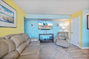 A living room with a beige sofa, a recliner, a TV on the wall, and a patterned rug. - Cab "Anna" Bay at Tiburon 5C by Seaside Vacations & Sales (Ocean City)