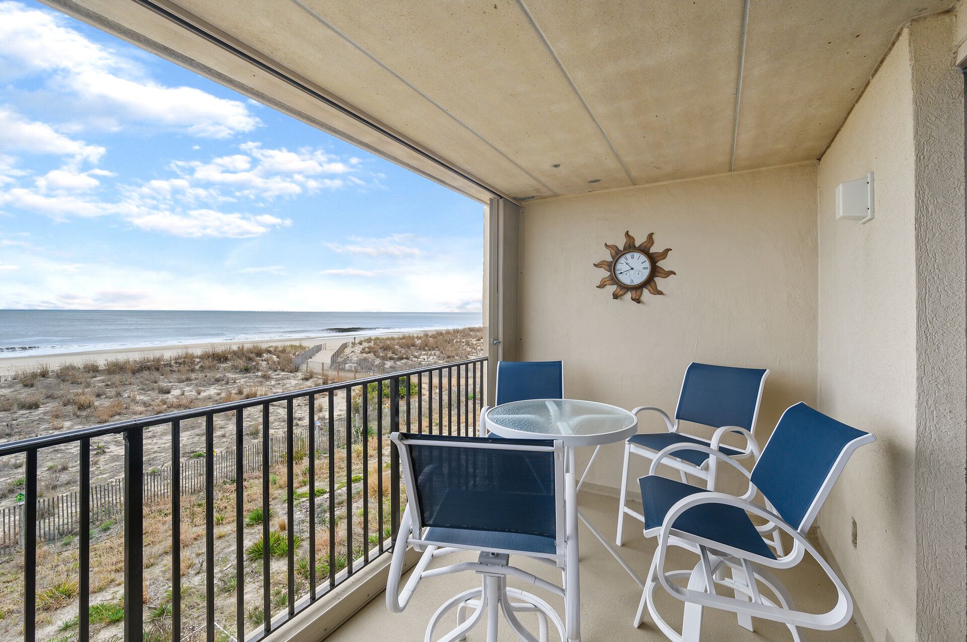 A balcony with a view of the beach, a round table, and two blue chairs.