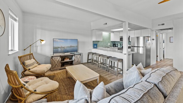 Ein modernes Wohnzimmer mit einer Couch, Sesseln, einem Couchtisch und einem Fernseher. Es gibt eine Küchenzeile mit Kühlschrank, Herd und Barhocker.