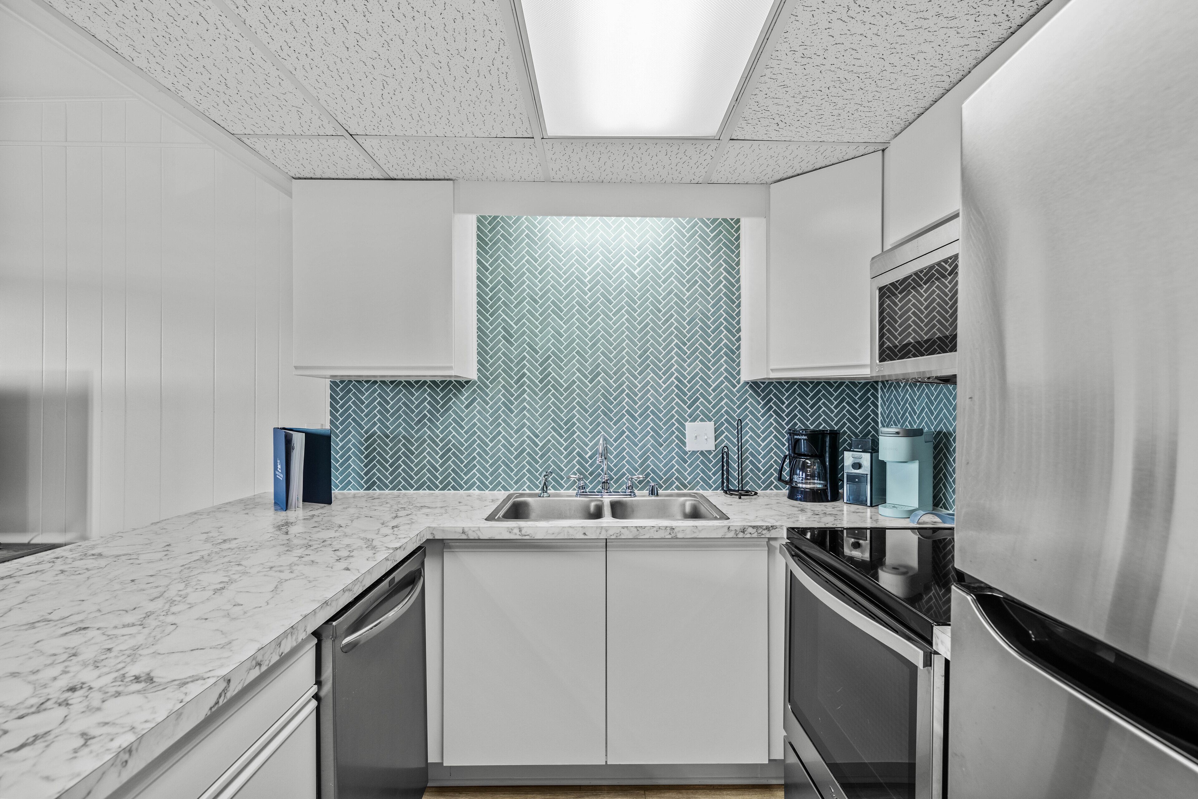 A modern kitchen with white cabinets, a marble countertop, a stainless steel refrigerator, and a herringbone-patterned backsplash.