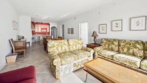 A spacious living area with a sofa, a red armchair, and a dining table. A kitchen with red cabinets is visible in the background. - Maresol 502 by Seaside Vacations & Sales (Ocean City)