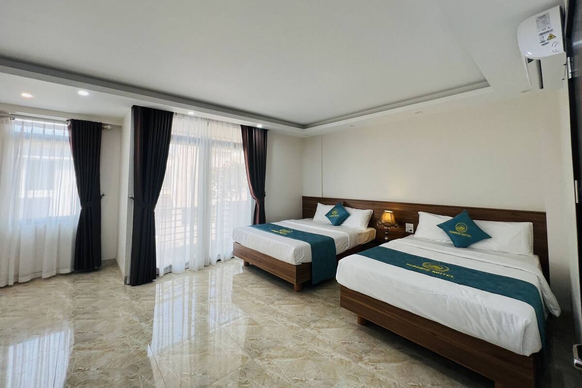 A hotel room with two beds, a large window with curtains, a marble floor, and a ceiling-mounted air conditioning unit.