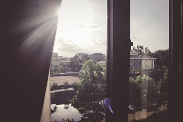A view through a window with sunlight streaming in, featuring a building, trees, and a body of water.