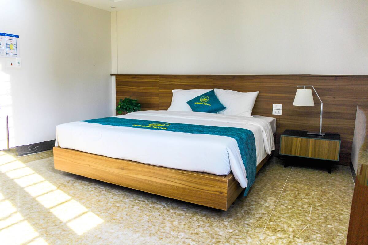A modern hotel room with a wooden bed, a nightstand with a lamp, and a view of the outside.