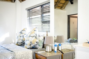 A neatly arranged hotel room with a bed, bedside tables, a lamp, and a window with blinds. - Nkanyi Wildlife House & Cottage (Hoedspruit)