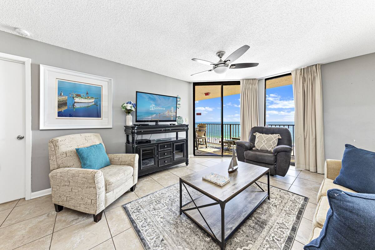 A living room with a television, a coffee table, a sofa, a chair, and a balcony with a view.