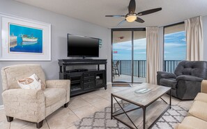 A living room with a television, a sofa, a chair, and a balcony with a view of the sea.